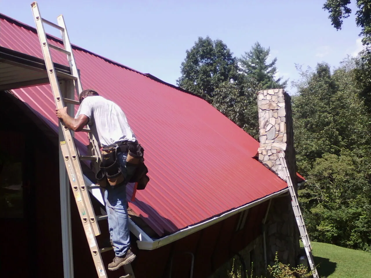 Roofer on a ladder beside a red metal roof and stone chimney — Soffit & Fascia Repair by Roofing Brothers in Langhorne